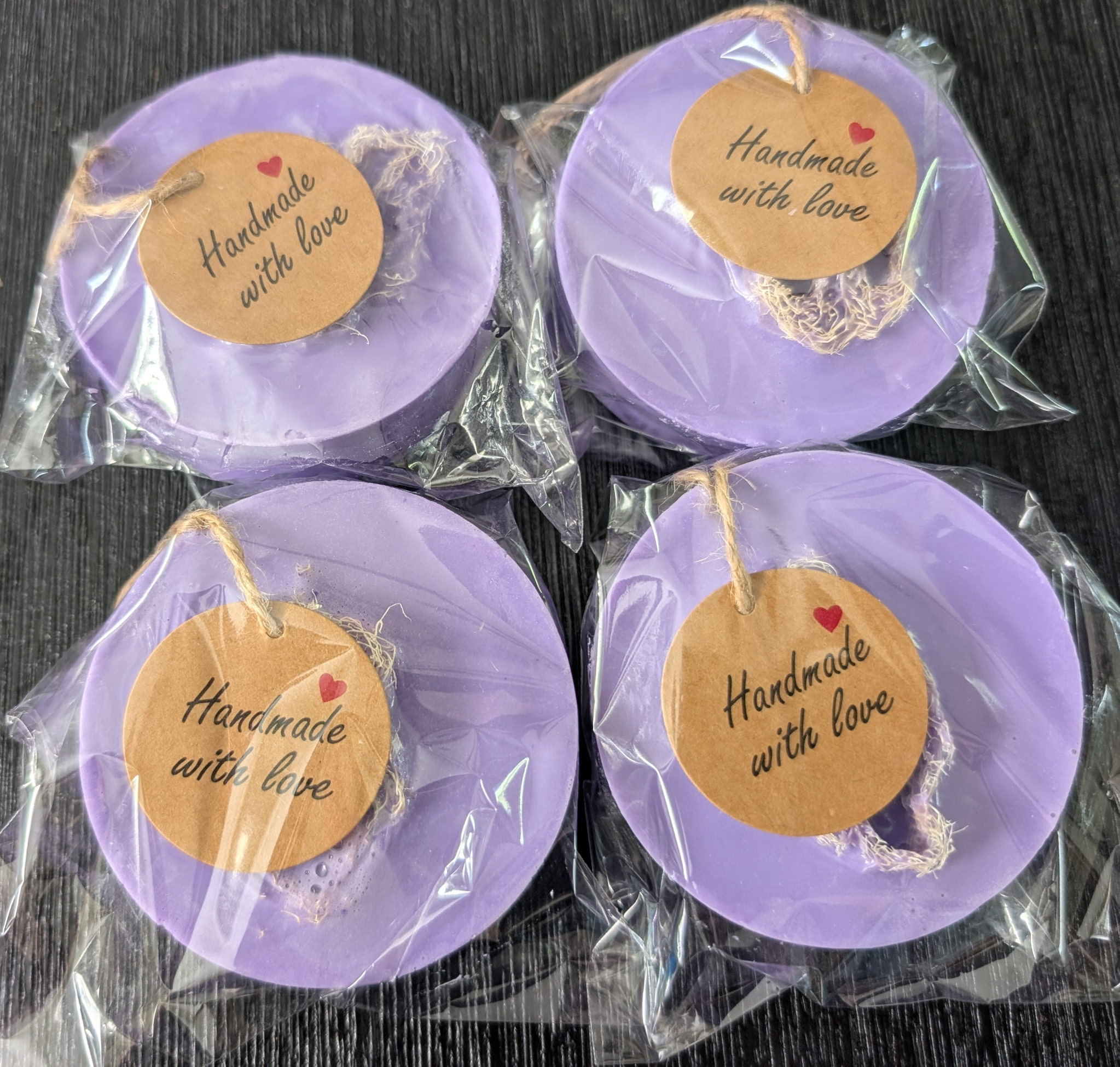 Four round, purple, handmade soap bars with embedded natural loofah, packaged in clear bags with 'Handmade with love' tags.